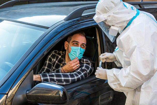 Man sitting in car, waiting for medical worker to perform drive-thru COVID-19 test, taking nasal swab sample through car window, PCR diagnostic for Coronavirus, doctor in PPE holding test kit. - Photo, Image