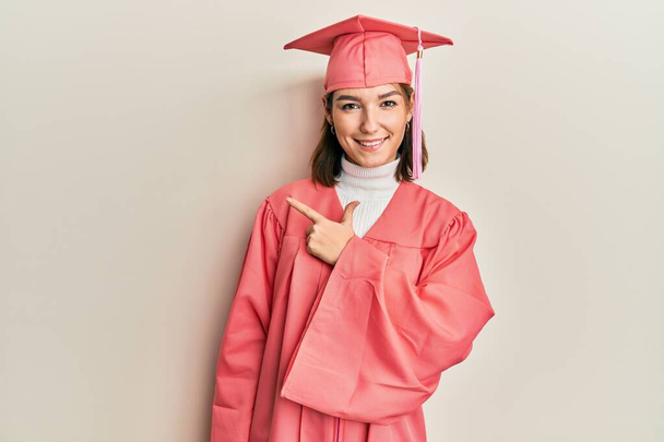Young caucasian woman wearing graduation cap and ceremony robe cheerful with a smile of face pointing with hand and finger up to the side with happy and natural expression on face  - Фото, изображение