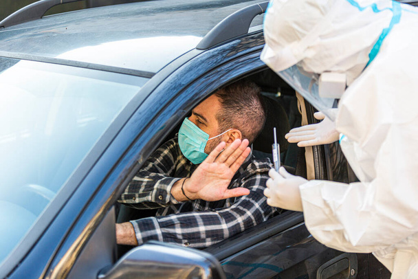 Mężczyzna odmawia pracownikowi medycznemu próby wykonania testu drive-thru COVID-19, pobrania próbki wymazu z nosa od pacjenta przez okno samochodu, diagnostyki PCR, lekarz w PPE posiadający zestaw testowy.  - Zdjęcie, obraz