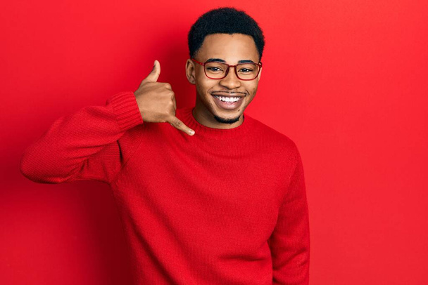 Jeune homme afro-américain portant des vêtements décontractés et des lunettes souriant faire geste de téléphone avec la main et les doigts comme parler au téléphone. communiquer les concepts.  - Photo, image