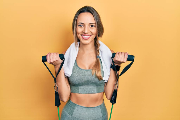 Schöne hispanische Frau, die den Armwiderstand trainiert, mit elastischen Armbändern, die mit einem fröhlichen und kühlen Lächeln im Gesicht lächeln. Zähne zeigen.  - Foto, Bild
