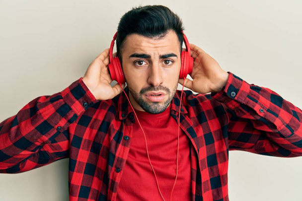 Joven hombre hispano escuchando música usando auriculares sin pistas y expresión confusa. concepto de duda.  - Foto, imagen