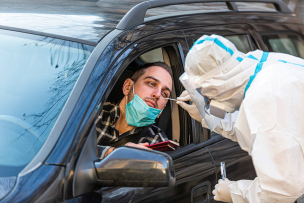 Pracownik medyczny wykonujący test drive-thru COVID-19, pobierający wymaz z nosa od pacjenta przez okno samochodu, diagnostykę PCR, lekarz w apteczce PPE. Trzyma paszporty.. - Zdjęcie, obraz