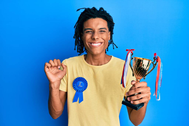 Young african american man wearing first place badge holding trophy screaming proud, celebrating victory and success very excited with raised arm  - Photo, image