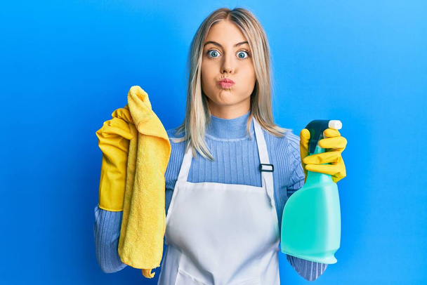 Beautiful blonde woman wearing cleaner apron holding cleaning products puffing cheeks with funny face. mouth inflated with air, catching air.  - Photo, Image
