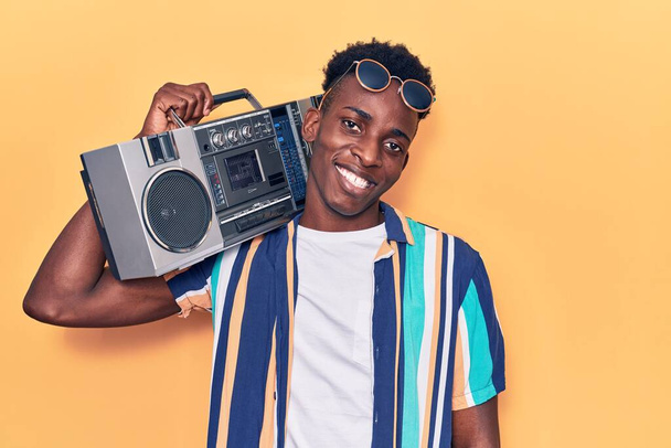Giovane uomo afroamericano che tiene in mano il boombox, ascolta musica che sembra positiva e felice in piedi e sorride con un sorriso fiducioso mostrando i denti  - Foto, immagini