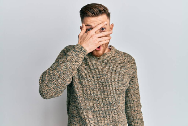 Young redhead man wearing casual winter sweater peeking in shock covering face and eyes with hand, looking through fingers afraid  - Foto, immagini
