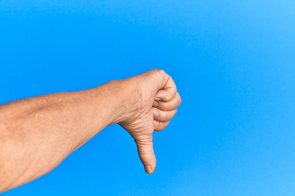 Ruka staršího hispánského muže nad modrým izolovaným pozadím dělá palce dolů odmítavé gesto, nesouhlas nelibost a negativní znamení  - Fotografie, Obrázek