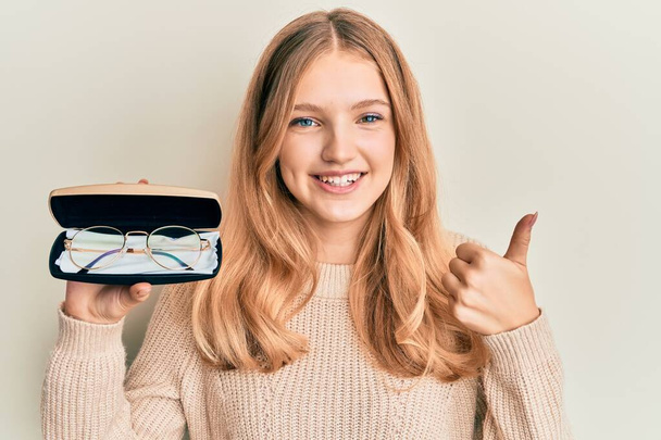 Belle jeune fille caucasienne tenant des lunettes dans un étui à lunettes souriant heureux et positif, pouce levé faisant excellent et signe d'approbation  - Photo, image