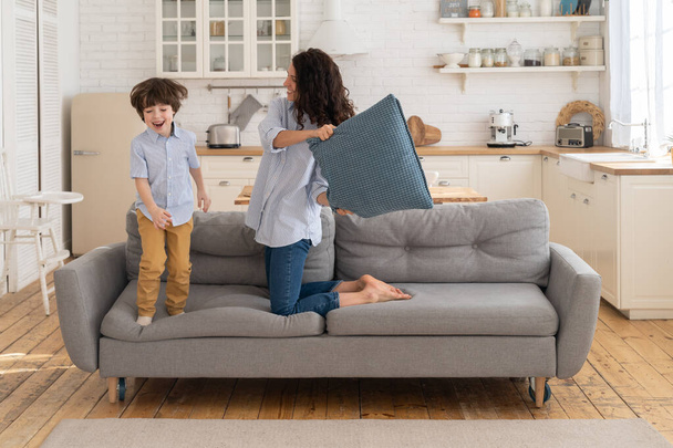 Mom and son having fun in living room fighting with pillows together. Family leisure and recreation time - Фото, изображение