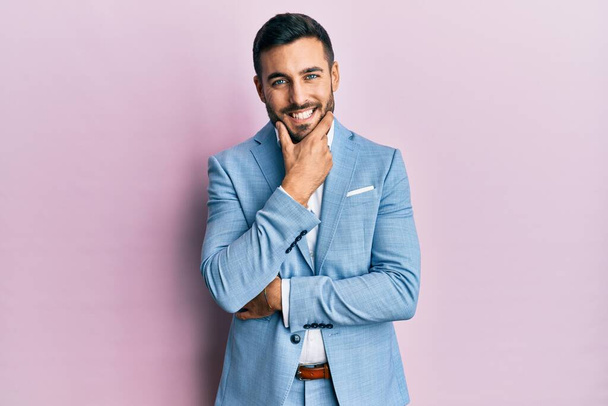 Der junge hispanische Geschäftsmann in Businessjacke blickt selbstbewusst in die Kamera und lächelt mit verschränkten Armen und erhobener Hand am Kinn. Positives Denken.  - Foto, Bild
