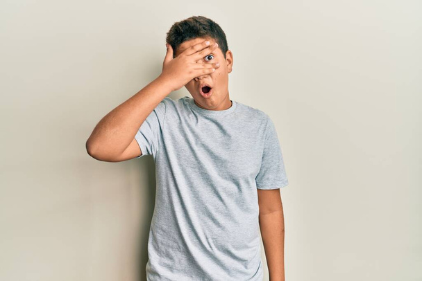 Teenager hispánský chlapec v neformální šedé tričko vykukující v šoku zakrývající obličej a oči rukou, dívající se skrz prsty s rozpačitým výrazem.  - Fotografie, Obrázek