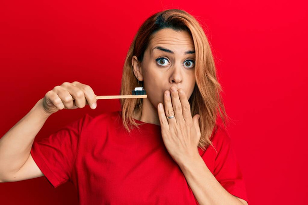 Іспанська молода жінка тримає зубну щітку з зубною пастою, закриваючи рот рукою, шокована і налякана за помилку. здивований вираз  - Фото, зображення