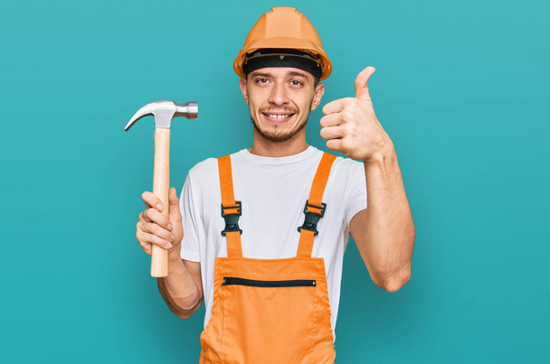 Hispanic young man wearing hardhat holding hammer smiling happy and positive, thumb up doing excellent and approval sign  - Фото, зображення