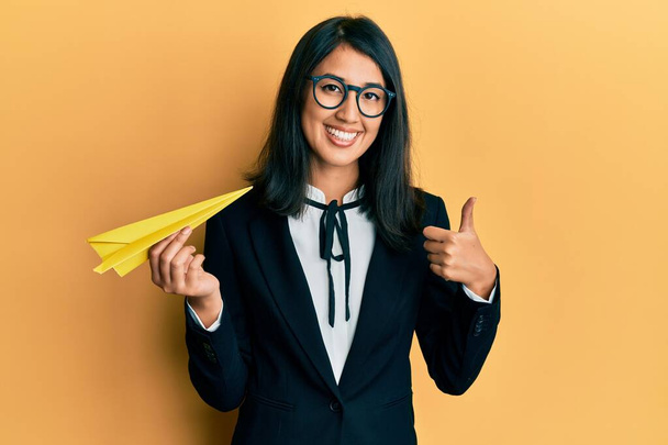 Hermosa asiática joven mujer de negocios sosteniendo avión de papel para el viaje de trabajo sonriendo feliz y positivo, pulgar hacia arriba haciendo excelente y aprobación signo  - Foto, imagen