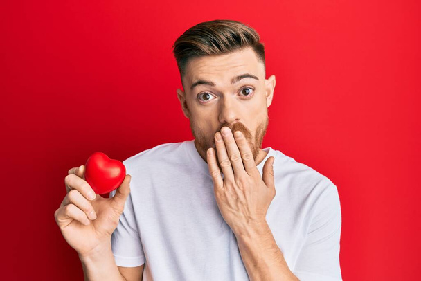 Young redhead man holding heart covering mouth with hand, shocked and afraid for mistake. surprised expression  - Foto, afbeelding