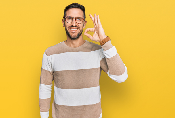 Handsome man with beard wearing casual clothes and glasses smiling positive doing ok sign with hand and fingers. successful expression.  - Photo, Image