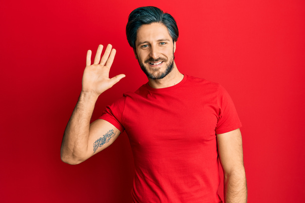 Joven hombre hispano con camiseta roja casual renunciando a decir hola feliz y sonriente, gesto de bienvenida amistoso  - Foto, imagen