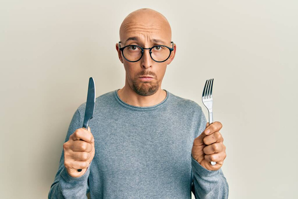 Hombre calvo con barba sosteniendo tenedor y cuchillo listo para comer deprimido y preocuparse por la angustia, llorando enojado y asustado. expresión triste.  - Foto, Imagen