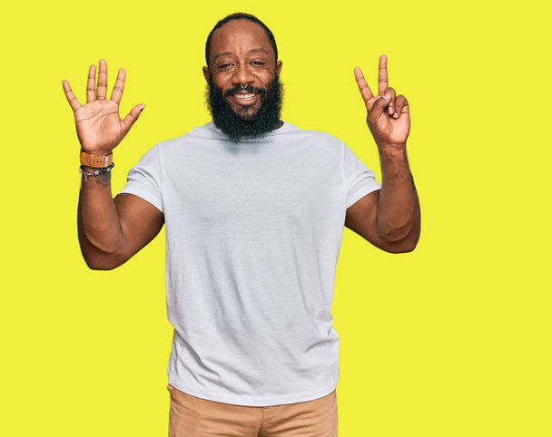 Joven hombre afroamericano vistiendo camiseta blanca casual mostrando y señalando con los dedos número siete mientras sonríe confiado y feliz.  - Foto, Imagen