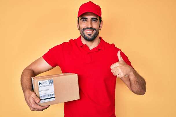 Hombre hispano guapo con barba sosteniendo paquete de entrega sonriendo feliz y positivo, pulgar hacia arriba haciendo excelente y signo de aprobación  - Foto, Imagen