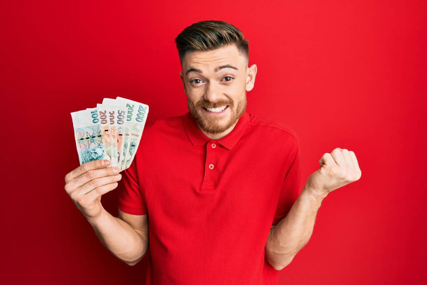 Junger rothaariger Mann mit tschechischen Kronen-Banknoten schreit stolz, feiert Sieg und Erfolg sehr aufgeregt mit erhobenem Arm  - Foto, Bild