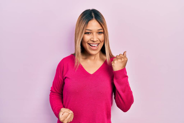 Красивая латиноамериканка в розовом свитере радуется и удивляется успеху с поднятыми руками и закрытыми глазами.  - Фото, изображение