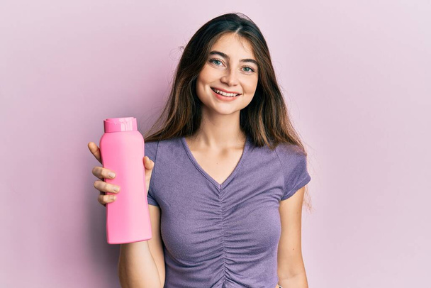 Молодая белая женщина с бутылкой шампуня выглядит позитивно и счастливо стоя и улыбаясь с уверенной улыбкой, показывая зубы  - Фото, изображение