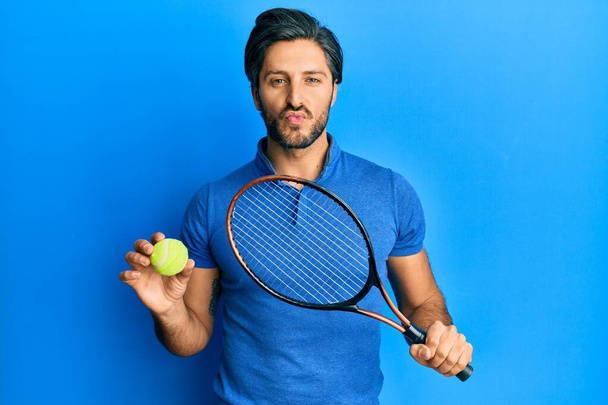 Молодой латиноамериканец играет в теннис, держа ракетку и мяч, глядя в камеру и дуя в поцелуй, будучи милым и сексуальным. выражение любви.  - Фото, изображение