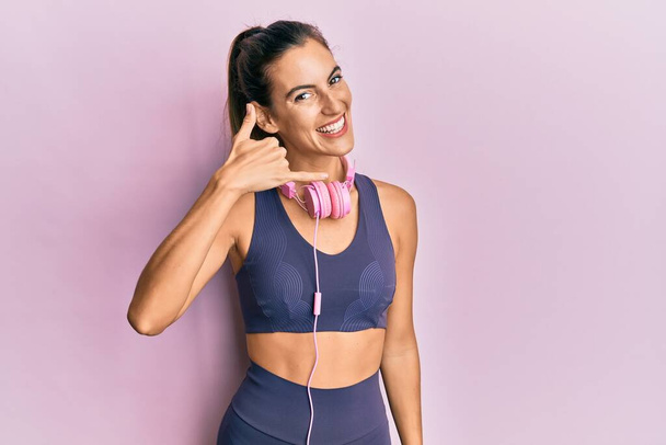 Young beautiful woman wearing gym clothes and using headphones smiling doing phone gesture with hand and fingers like talking on the telephone. communicating concepts.  - Foto, Imagem