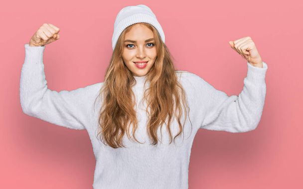 Joven chica caucásica con suéter de lana y gorra de invierno mostrando los músculos de los brazos sonriendo orgulloso. concepto de fitness.  - Foto, Imagen