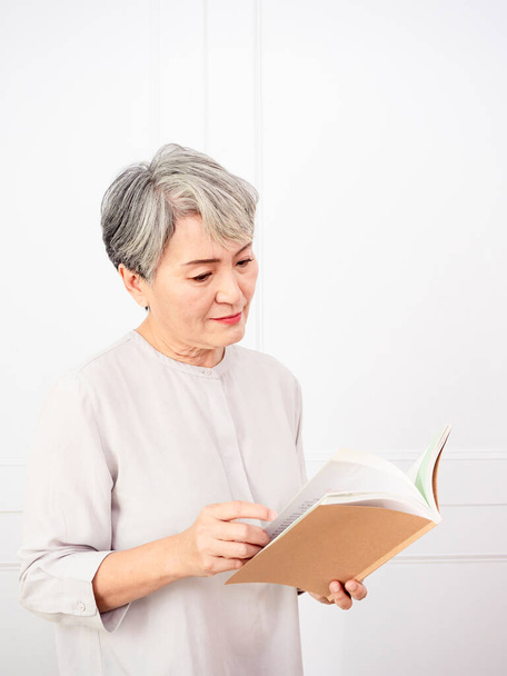 Senior gris pelo asiático mujer celebración y lectura libro en casa. - Foto, imagen