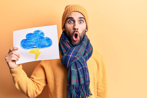 Mladý pohledný muž s vousy v zimním oblečení drží hrom čerpat strach a ohromen s otevřenými ústy pro překvapení, nevěřící tvář  - Fotografie, Obrázek