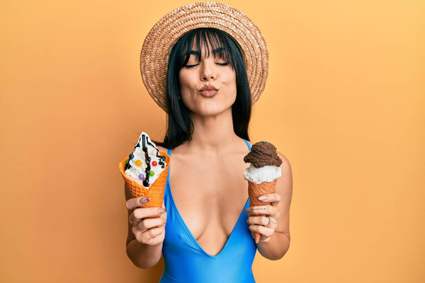 Young brunette woman with bangs wearing swimwear holding two ice cream cones looking at the camera blowing a kiss being lovely and sexy. love expression.  - Photo, image