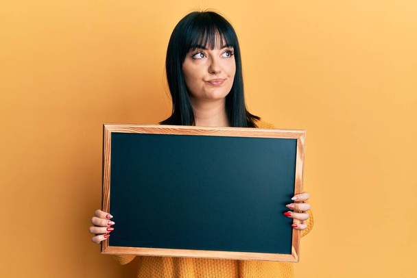 Young hispanic woman holding blackboard smiling looking to the side and staring away thinking.  - Valokuva, kuva