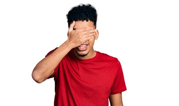 Jeune homme afro-américain portant un t-shirt rouge décontracté souriant et riant avec la main sur le visage couvrant les yeux pour la surprise. concept aveugle.  - Photo, image