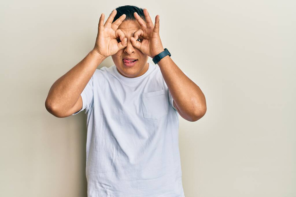 Young latin man wearing casual white t shirt doing ok gesture like binoculars sticking tongue out, eyes looking through fingers. crazy expression.  - Foto, imagen