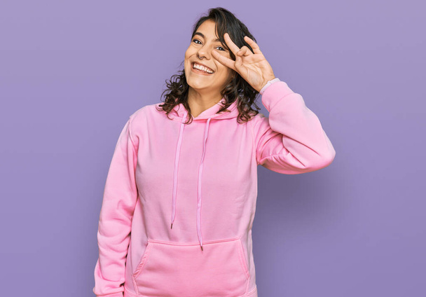 Junge hispanische Frau in lässigem Sweatshirt, die Friedenssymbol mit den Fingern über dem Gesicht trägt, fröhlich lächelnd den Sieg zeigend  - Foto, Bild