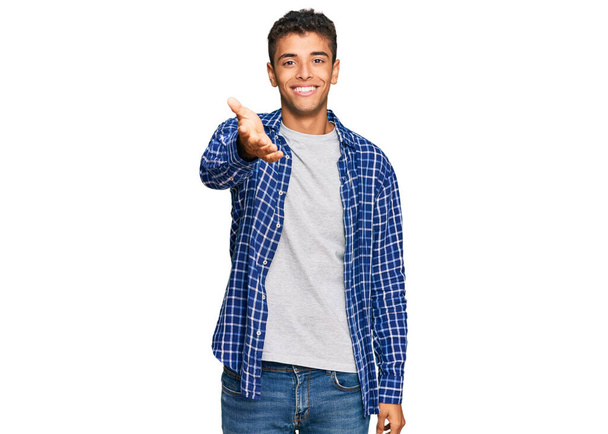 Joven hombre afroamericano guapo vistiendo ropa casual sonriendo amistoso ofreciendo apretón de manos como saludo y bienvenida. negocios exitosos.  - Foto, Imagen