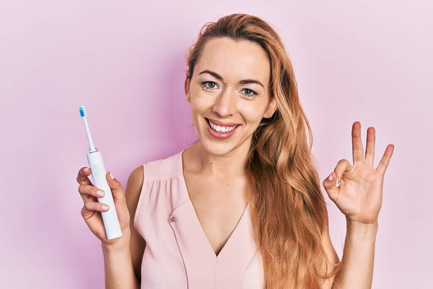 Elinde elektrikli diş fırçası olan beyaz bir kadın parmaklarıyla işaret veriyor, gülümseyerek mükemmel bir sembol oluşturuyor.  - Fotoğraf, Görsel