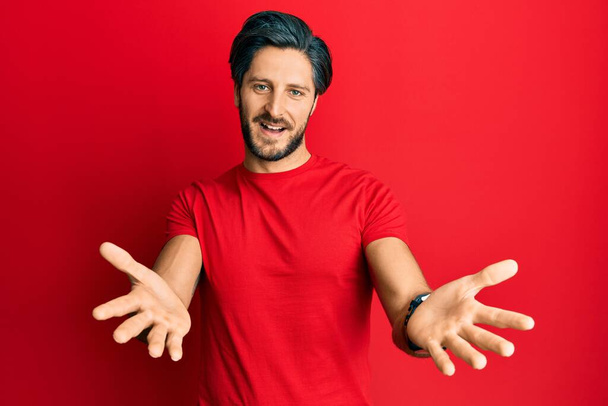 Young hispanic man wearing casual red t shirt smiling cheerful offering hands giving assistance and acceptance.  - 写真・画像