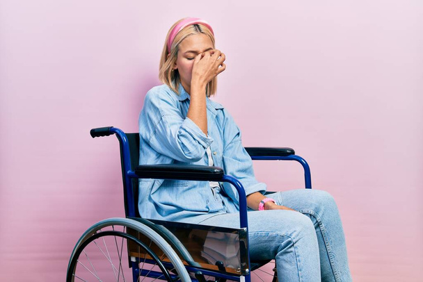 Schöne blonde Frau sitzt im Rollstuhl und reibt sich müde Nase und Augen und spürt Müdigkeit und Kopfschmerzen. Konzept für Stress und Frustration.  - Foto, Bild