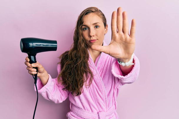 Mujer rubia joven con albornoz usando secador de pelo con la mano abierta haciendo stop sign con expresión seria y segura, gesto de defensa  - Foto, imagen