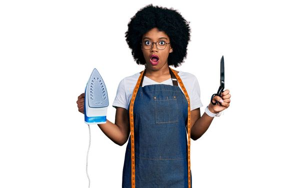 Mladá africká americká žena krejčí návrhářka v ateliéru zástěra drží železo a nůžky v šoku tvář, vypadá skeptický a sarkastický, překvapený s otevřenými ústy  - Fotografie, Obrázek