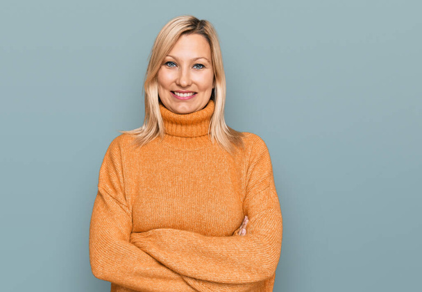 Biała kobieta w średnim wieku, ubrana w luźny zimowy sweter, uśmiechnięta z skrzyżowanymi ramionami patrzącymi w kamerę. osoba pozytywna.  - Zdjęcie, obraz