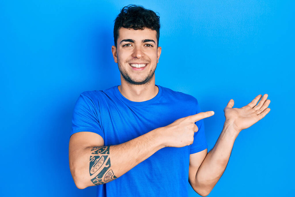 Jeune homme hispanique portant t-shirt bleu décontracté étonné et souriant à la caméra tout en présentant avec la main et pointant du doigt.  - Photo, image