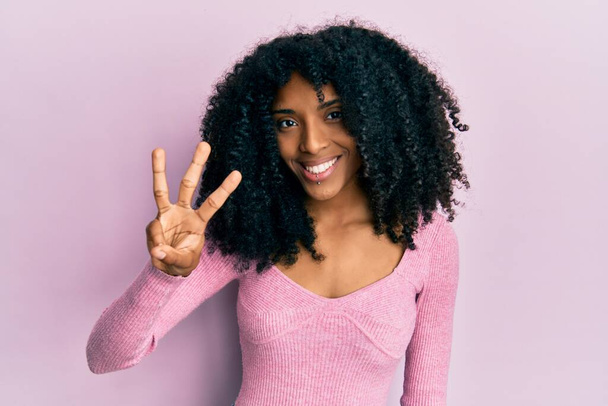 Африканская американка с афроволосами в розовой рубашке, показывающая пальцами номер три, улыбаясь уверенно и счастливо.  - Фото, изображение