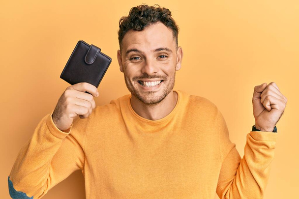 Young hispanic man holding leather wallet screaming proud, celebrating victory and success very excited with raised arm  - Fotografie, Obrázek