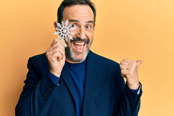 Middle age hispanic man holding snowflake over eye pointing thumb up to the side smiling happy with open mouth  - 写真・画像