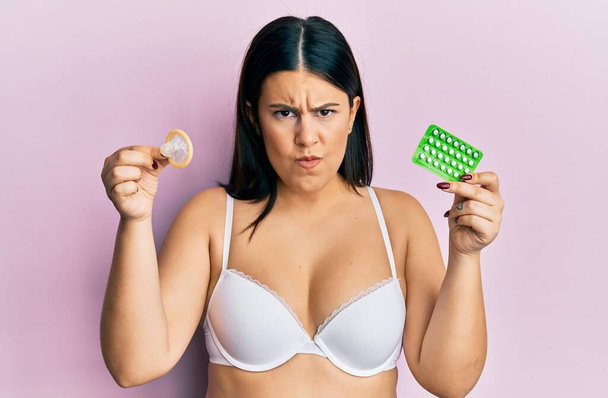 Krásná brunetka žena drží kondom a antikoncepci pilulky skeptické a nervózní, zamračený rozrušený kvůli problému. negativní osoba.  - Fotografie, Obrázek